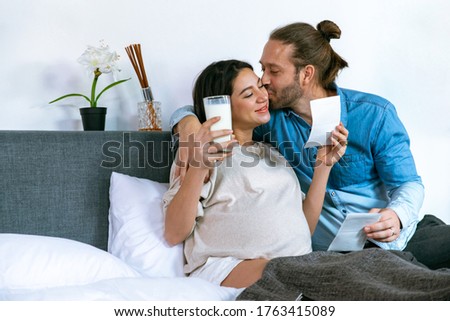 Smiling young beautiful Caucasian pregnant woman wife drinking fresh milk and looking ultrasound baby picture together with her husband on the bed. Family relationship and maternity healthcare concept