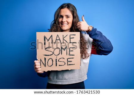 Beautiful activist woman holding banner with make some noise message over blue background happy with big smile doing ok sign, thumb up with fingers, excellent sign