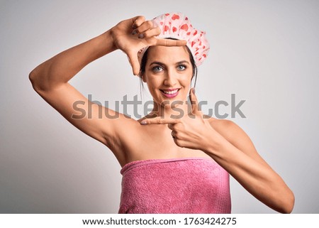 Young brunette woman with blue eyes wearing bath towel and shower cap smiling making frame with hands and fingers with happy face. Creativity and photography concept.