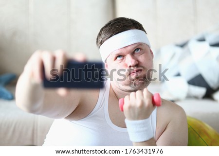 Young man in sport headband doing exercise with dumbbell and trying not to breath while taking picture with smartphone