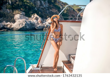 Beautiful female model happy with holiday trip on a luxury yacht. She wearing bikini on slim body, sunglasses and hat. She enjoying sunshine and sea view, standing in relaxed pose on the yacht deck.  