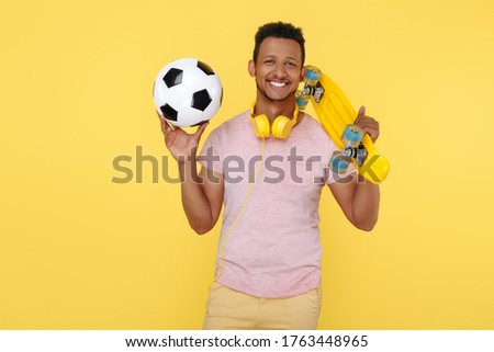 Gleeful african american young man wearing headphones holding soccer ball and skateboard over yellow background.