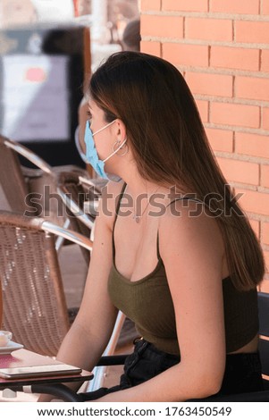 Close up of a young woman using a surgical mask and sitting at an outdoor terrace looking away from the camera on an out of focus background. Safety and new lifestyle concept.