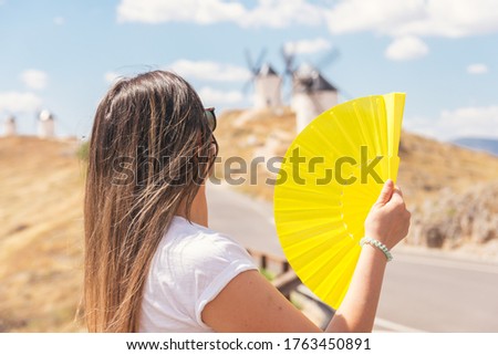 Blonde girl with a yellow fan in her hand fanning herself. Traditional windmills in the background.