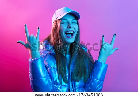 Happy excited woman showing rock gesture with fingers, visiting cool party, yelling something happily, wearing baseball cap and jacket, poses against pink neon wall.