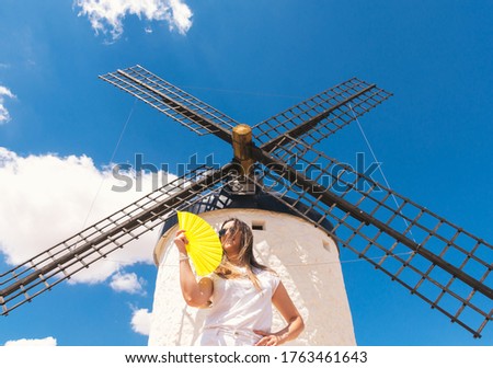 Blonde girl with a yellow fan in her hand fanning herself. Traditional windmill background and blue sky