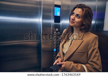 Cute brunette female in trendy coat staying in lift alone with mobile in hands and smiling Royalty-Free Stock Photo #1763463569