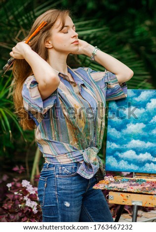 Young beautiful girl 
in work clothes draws a picture while with an easel 
in the botanical garden. Girl enjoy solitude with nature.