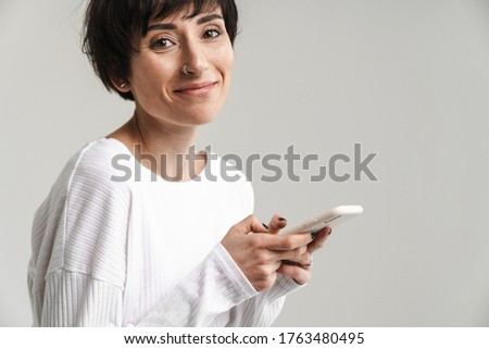 Portrait of an attractive smiling short brunette haired woman wearing sweater standing isolated over white background, using mobile phone