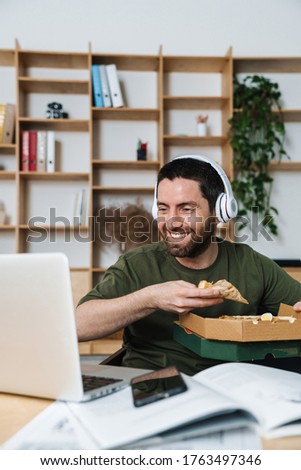 Photo of smiling bearded man in headphones eating pizza while working with laptop in office