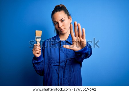 Young beautiful painter woman with blue eyes painting wearing uniform using paint brush with open hand doing stop sign with serious and confident expression, defense gesture