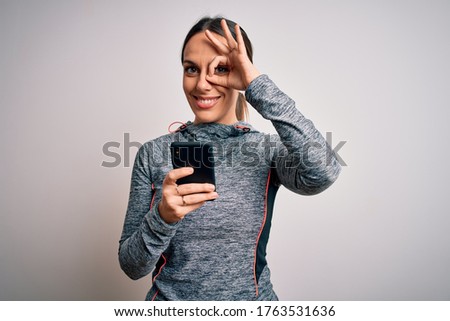 Young fitness woman wearing sport workout clothes using smartphone app with happy face smiling doing ok sign with hand on eye looking through fingers