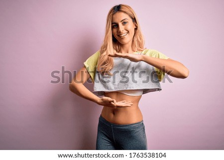 Young beautiful blonde sportswoman doing sport wearing sportswear over pink background gesturing with hands showing big and large size sign, measure symbol. Smiling looking at the camera. Measuring.