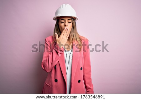 Young beautiful brunette architect woman wearing safety helmet over pink background bored yawning tired covering mouth with hand. Restless and sleepiness.
