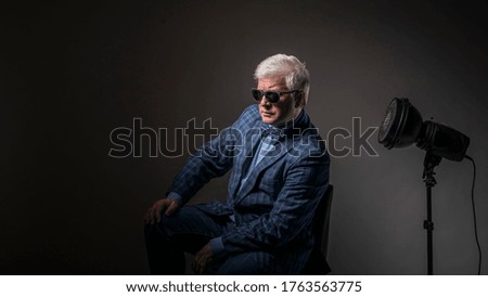 Portrait of a courageous and brutal, majestic, rich and mature man, financier, businessman in black glasses with gray hair, in a blue jacket and bow tie on a gray background