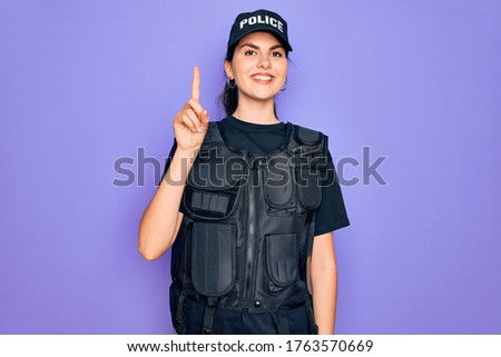 Young police woman wearing security bulletproof vest uniform over purple background showing and pointing up with finger number one while smiling confident and happy.