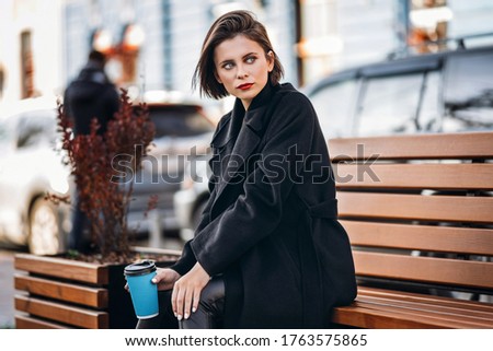Young woman with short haircut and red lips, dressed in black coat, sits on a bench in the city and holds a cup of coffee.