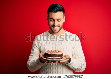 Young handsome man holding colorful birthday sweet cake over red isolated background with a happy face standing and smiling with a confident smile showing teeth
