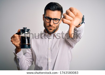 Young handsome man making coffee using french press coffeemaker over isolated background with angry face, negative sign showing dislike with thumbs down, rejection concept