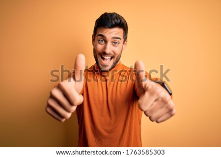 Young handsome man with beard wearing casual sweater standing over yellow background approving doing positive gesture with hand, thumbs up smiling and happy for success. Winner gesture. Royalty-Free Stock Photo #1763585303