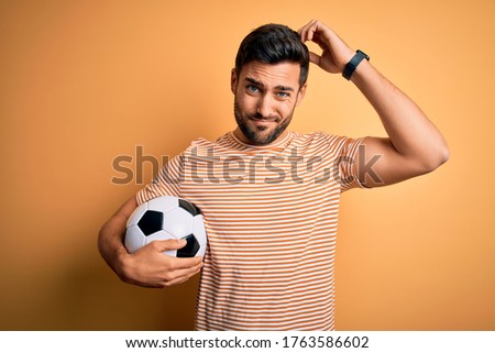 Handsome player man with beard playing soccer holding footballl ball over yellow background confuse and wondering about question. Uncertain with doubt, thinking with hand on head. Pensive concept.