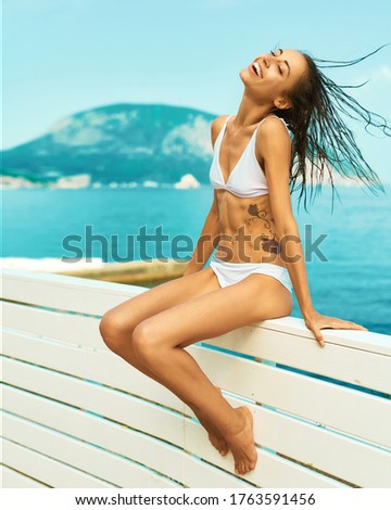 Happy tanned girl in white bikini with fit body posing flying hair, sunbathing on quay with mountain and sea on background. Bright summer vacation portrait