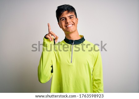 Young handsome sportsman doing sport wearing sportswear over isolated white background showing and pointing up with finger number one while smiling confident and happy.