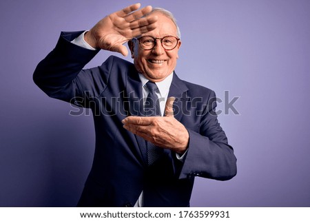 Grey haired senior business man wearing glasses and elegant suit and tie over purple background smiling making frame with hands and fingers with happy face. Creativity and photography concept.