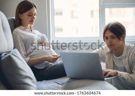 Pleased pregnant lady looking at the baby scan picture in her hands and smiling while her husband working on the laptop