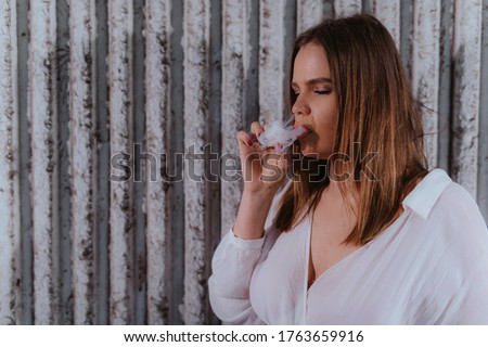 A beautiful young caucasian girl in a white shirt stands in front of a ribbed wall and smokes a cigar
