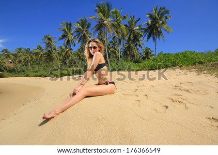 Beautiful girl in a bathing suit and sunglasses sitting on sand near the ocean under the palm trees