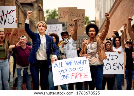 Multi-ethnic crowd of people with raised fists protesting for human rights while carrying anti-racism placards through city streets.  Royalty-Free Stock Photo #1763678525
