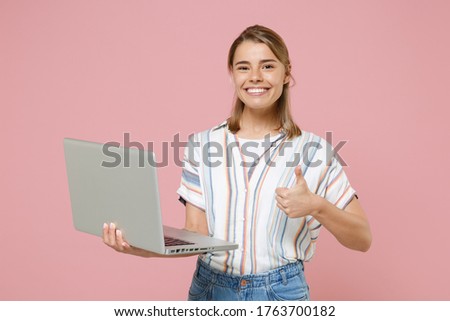 Smiling young blonde woman girl in casual striped shirt posing isolated on pastel pink background studio. People lifestyle concept. Mock up copy space. Working on laptop pc computer, showing thumb up