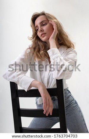 portrait of young pensive caucasian woman posing in shirt and blue jeans, sitting on black chair in white studio. model tests of pretty girl in basic clothes on cyclorama. attractive female poses