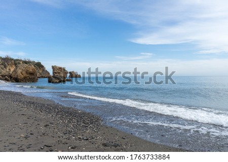 Big rocks in the blue sea with tiny waves and little foam, shells on a wet beach.