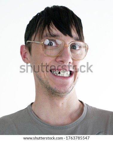 Close up portrait of Caucasian young man with greasy long black hair, big dark eyes, old glasses in t-shirt stupid smiling, show broken teeth, looking directly at camera isolated on white background. Royalty-Free Stock Photo #1763785547