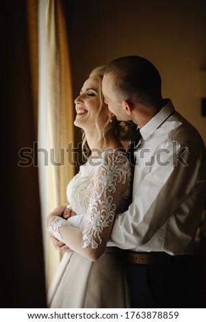 Bride and groom in a hotel room. The groom hugs and kisses the bride in the neck.
