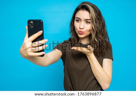 Young cheerful attractive woman taking selfie on the camera sending kiss of her phone isolated on the blue background.