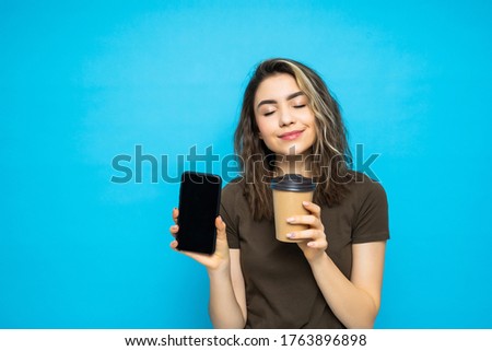 Young woman holding phone with screen for copy space and coffee to go isolated on blue background
