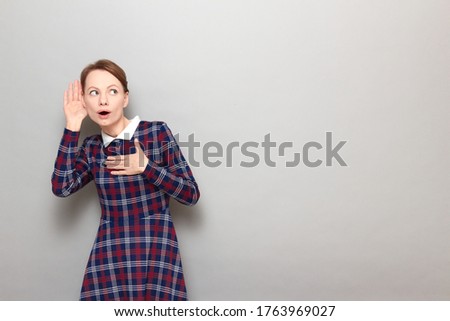 Studio portrait of curious girl wearing checkered dress, placing hand near ear, eavesdropping conversation, listening gossip, secrets or latest news, standing over gray background, with copy space