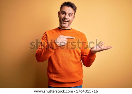 Young handsome man wearing orange casual sweater standing over isolated yellow background amazed and smiling to the camera while presenting with hand and pointing with finger.