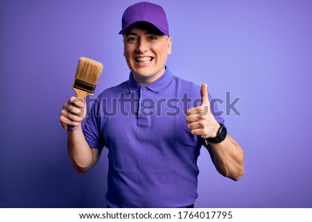 Young modern decorator painter man holding paint brush over purple background happy with big smile doing ok sign, thumb up with fingers, excellent sign