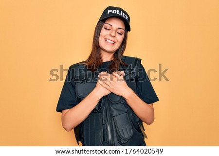 Young beautiful brunette policewoman wearing police uniform bulletproof and cap smiling with hands on chest with closed eyes and grateful gesture on face. Health concept.