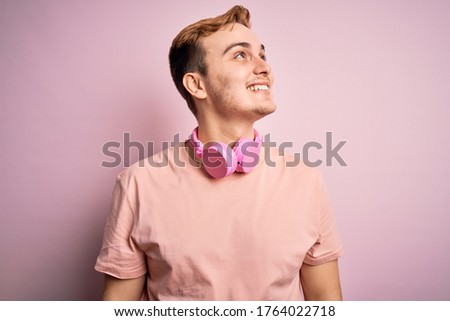 Young handsome redhead man listening to music using headphones over pink background looking away to side with smile on face, natural expression. Laughing confident.