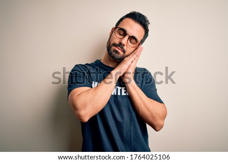 Handsome man with beard wearing t-shirt with volunteer message over white background sleeping tired dreaming and posing with hands together while smiling with closed eyes.