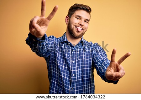 Young blond businessman with beard and blue eyes wearing shirt over yellow background smiling with tongue out showing fingers of both hands doing victory sign. Number two.
