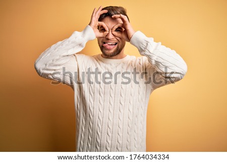 Young blond man with beard and blue eyes wearing white sweater over yellow background doing ok gesture like binoculars sticking tongue out, eyes looking through fingers. Crazy expression.