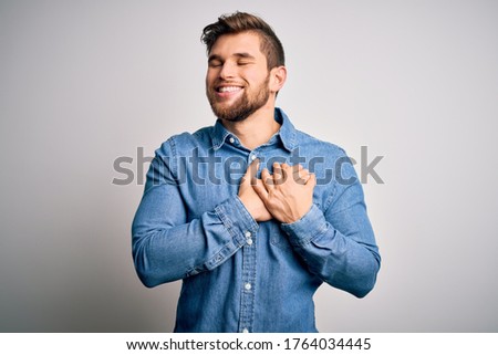 Young handsome blond man with beard and blue eyes wearing casual denim shirt smiling with hands on chest with closed eyes and grateful gesture on face. Health concept.