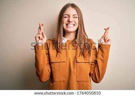 Young beautiful redhead woman wearing casual sweater over isolated white background gesturing finger crossed smiling with hope and eyes closed. Luck and superstitious concept.