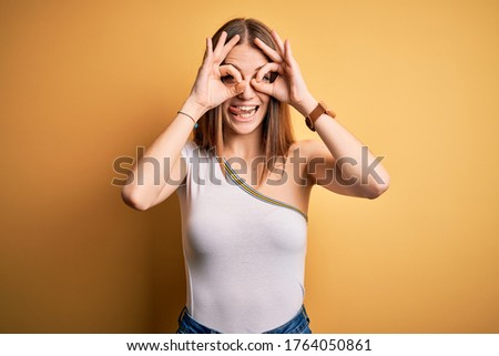Young beautiful redhead woman wearing casual t-shirt over isolated yellow background doing ok gesture like binoculars sticking tongue out, eyes looking through fingers. Crazy expression.